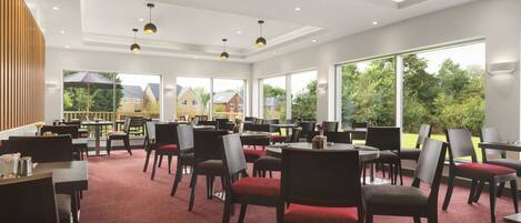 A spacious indoor dining area with multiple tables and chairs, large windows offering a view of the outside, and pendant lighting.