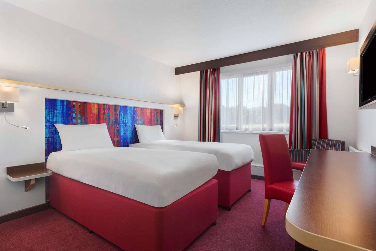 Ein Hotelzimmer mit zwei Betten, einem roten Sofa, einem Holztisch und einem farbenfrohen, abstrakten Gemälde an der Wand.