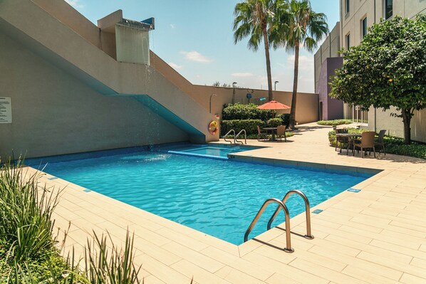 Una piscina con agua cristalina azul, rodeada por un solado de baldosas, una palmera y un edificio con toldo rojo.
