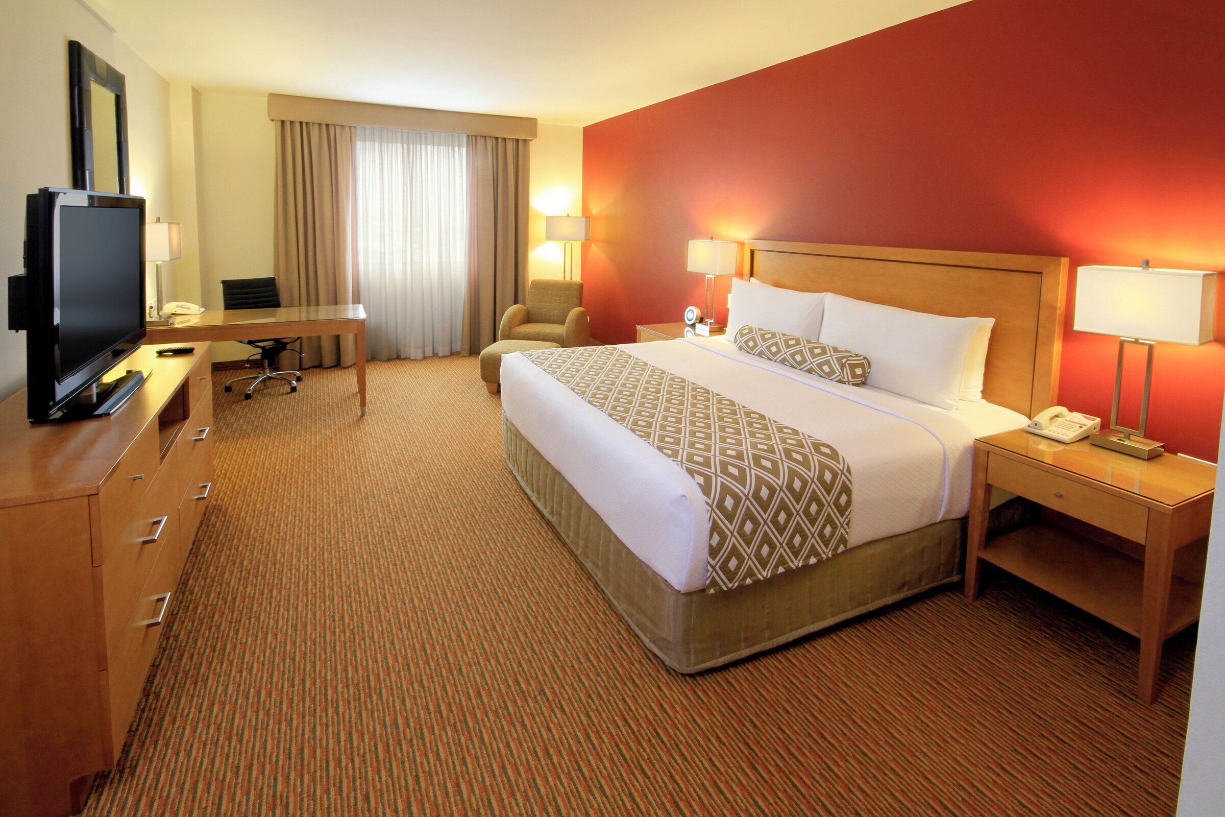 A hotel room with a bed, bedside tables, a desk with a chair, a television, and a large window with curtains.