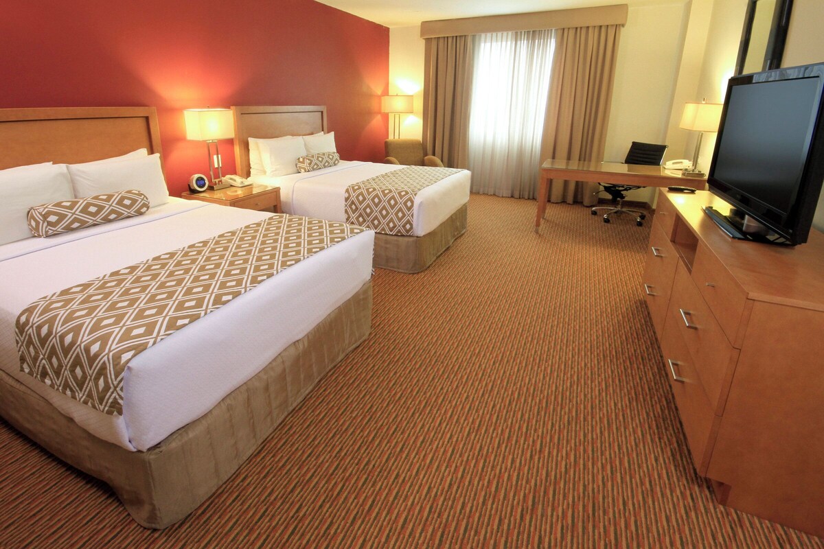 Quarto de hotel com duas camas, uma escrivaninha, televisão e janela com cortinas.