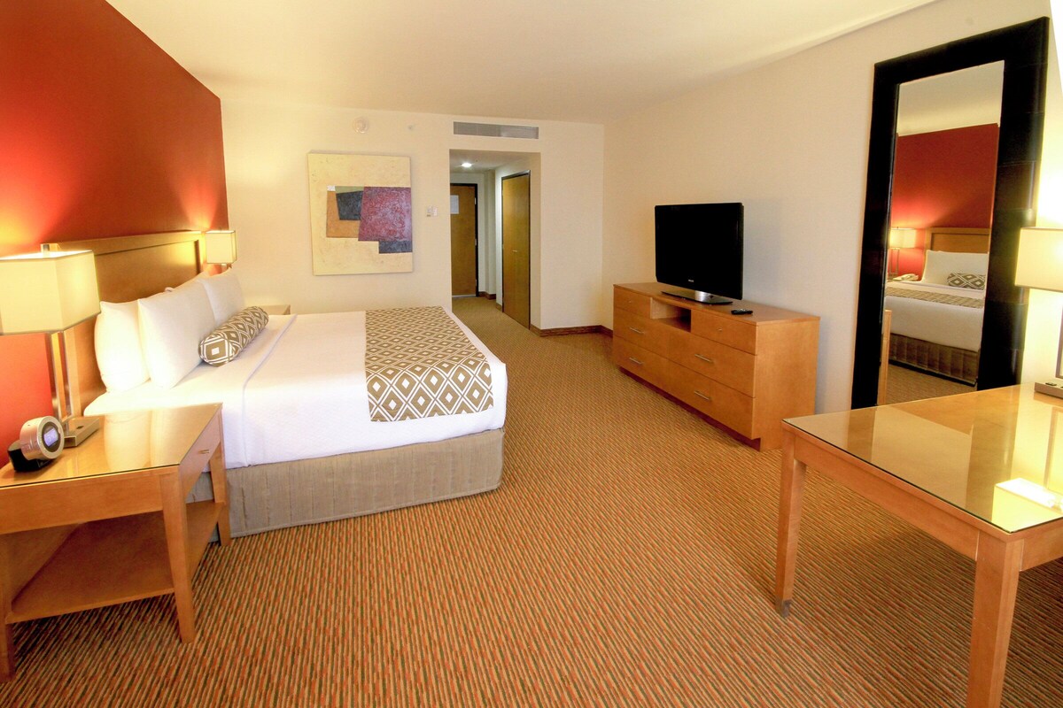 Quarto de hotel com cama, mesa de cabeceira, televisão, cômoda e espelho.