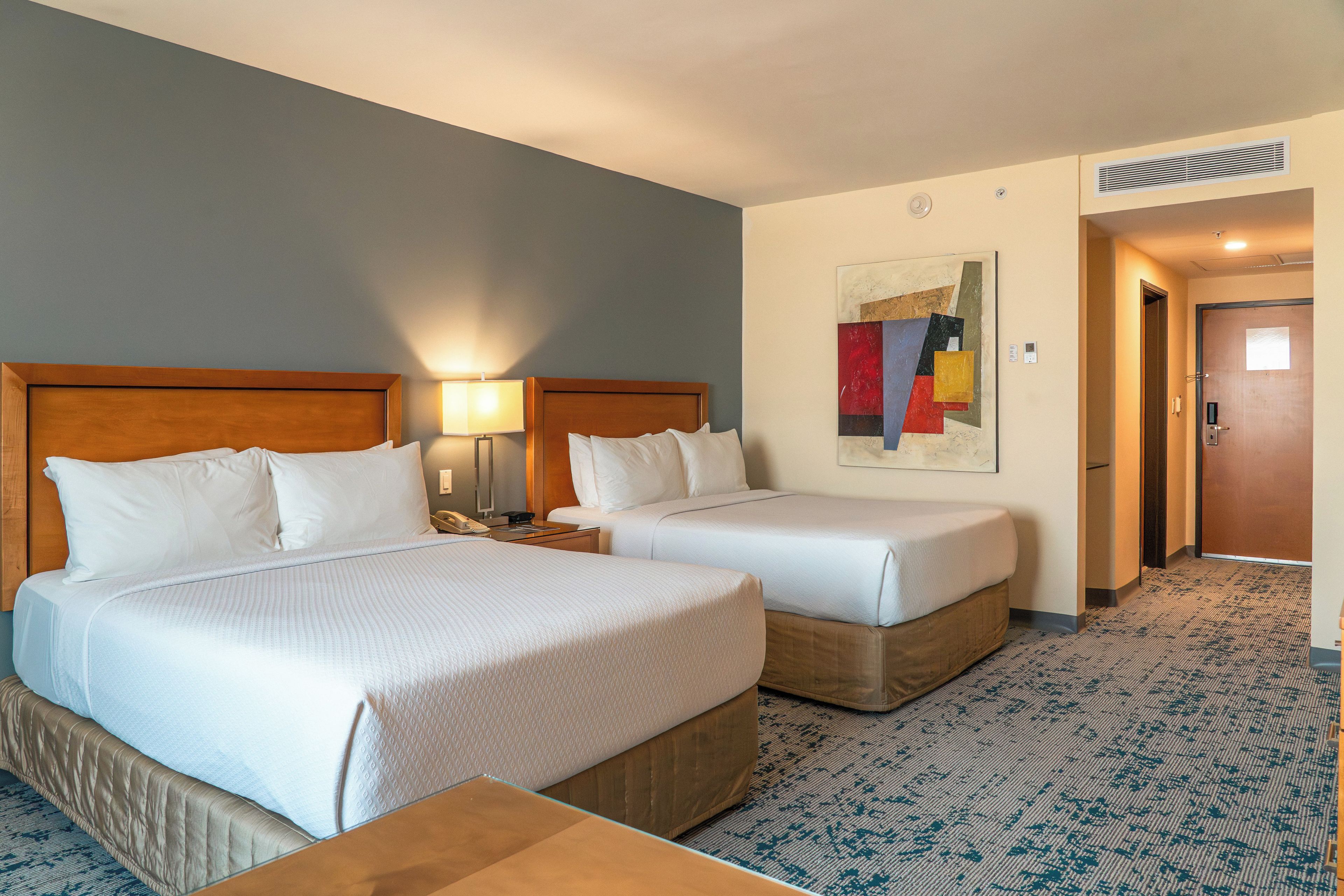 A hotel room with two beds, a nightstand, a lamp, a painting on the wall, and a door.