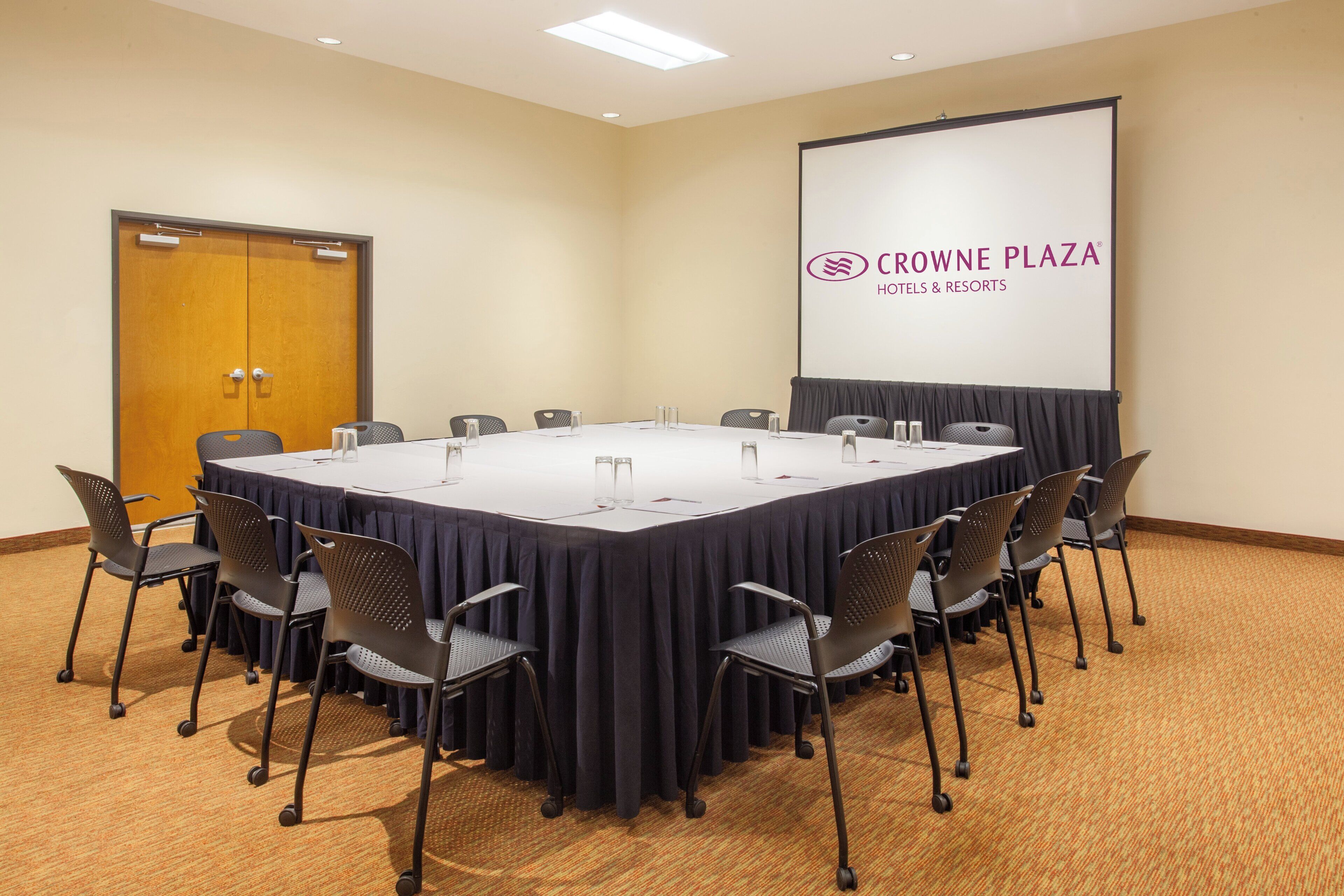 Un salón de conferencias con una larga mesa, sillas y una pantalla que muestra la marca 'CROWNE PLAZA HOTELS & RESORTS'.