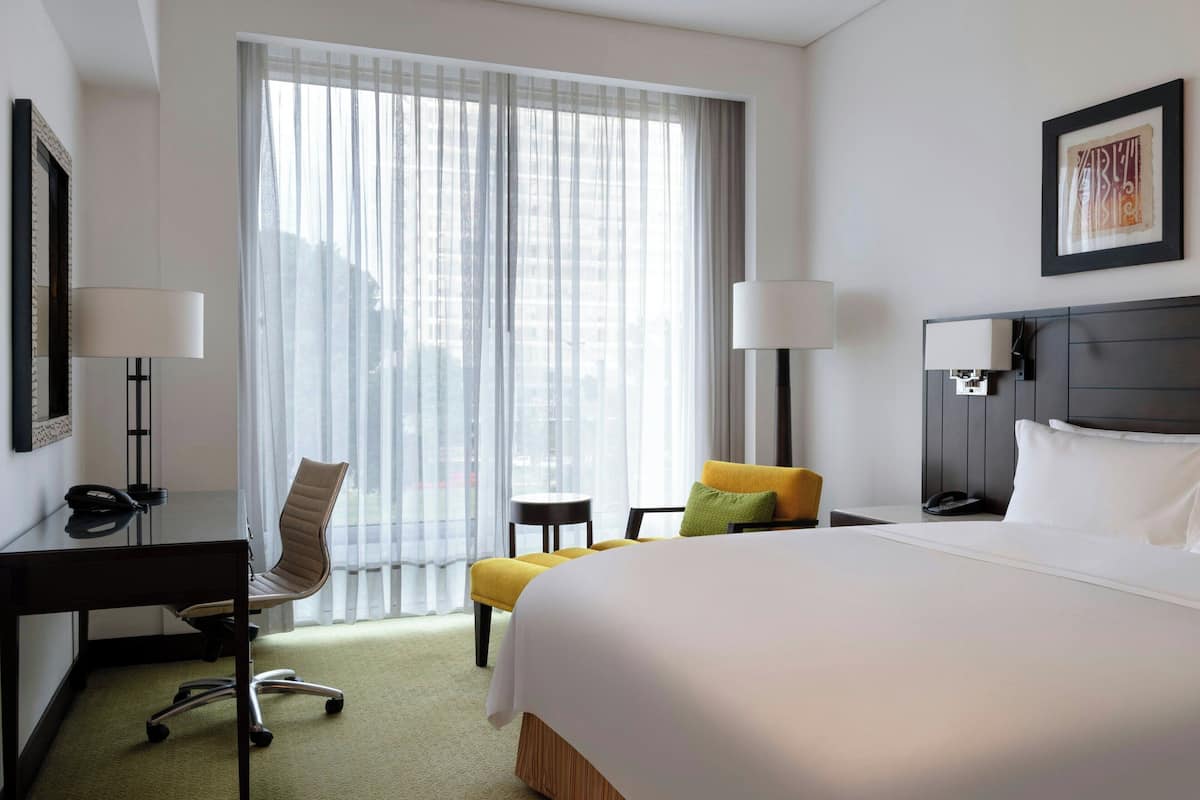 A hotel room with a large bed, a desk with a chair, a small table, and a window with sheer curtains.