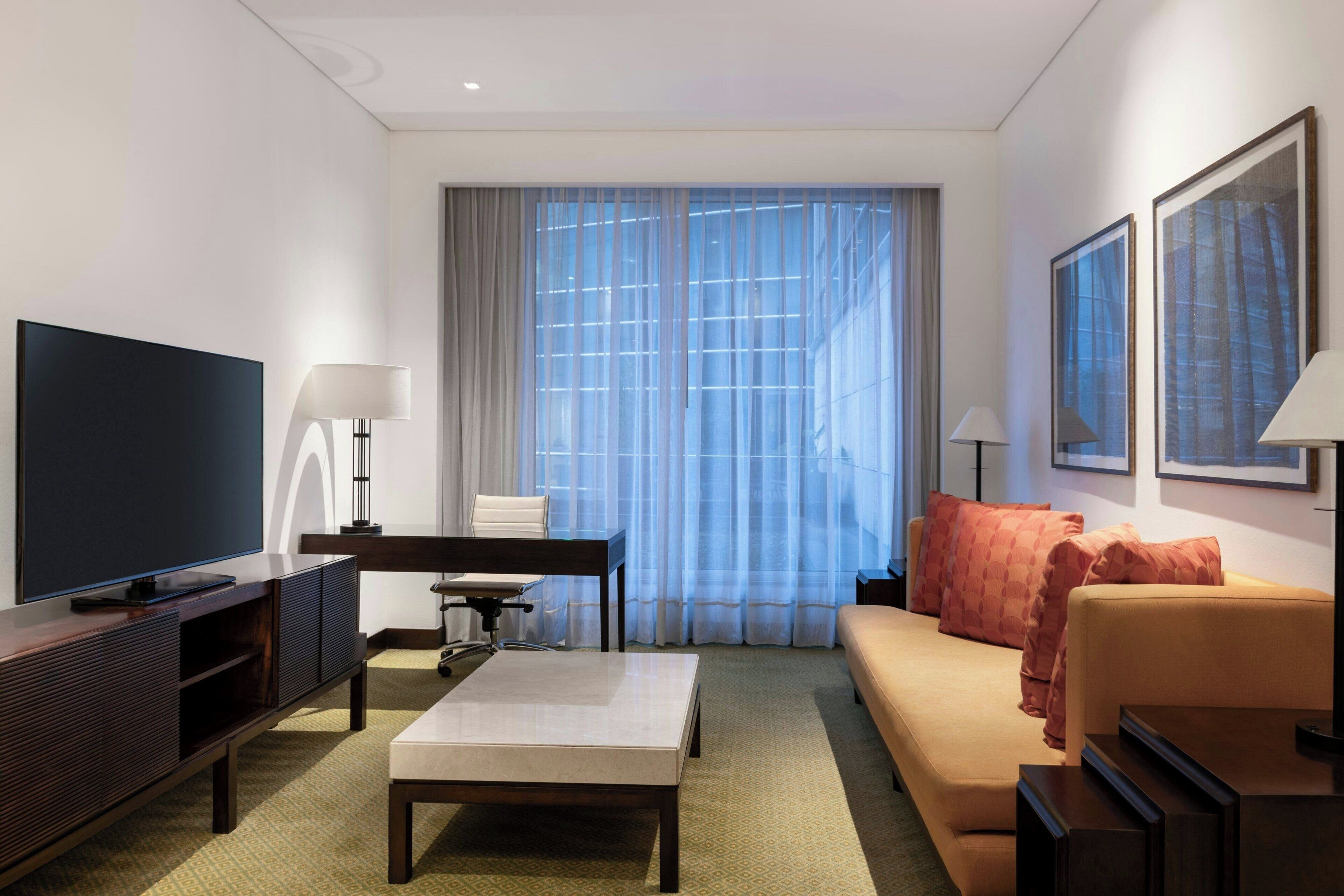 A modern hotel room with a flat-screen TV, a desk, a sofa, and a large window with sheer curtains.