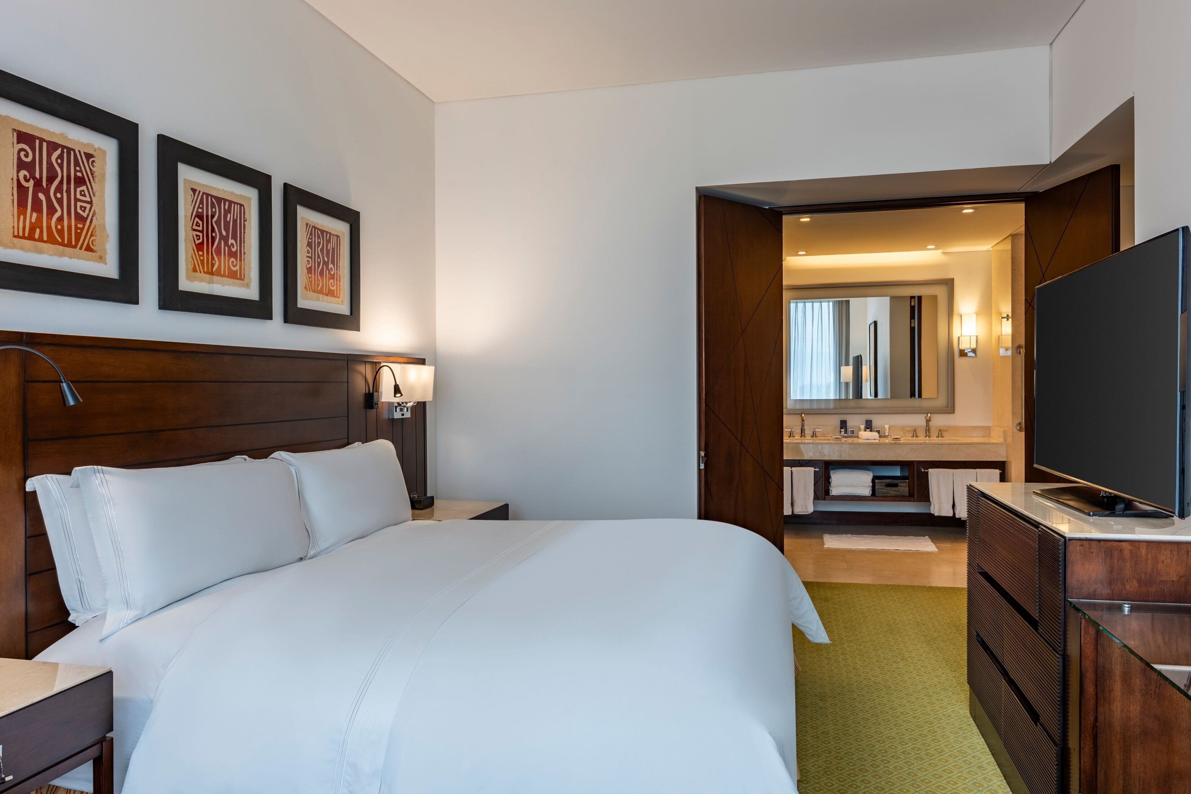 Ein Hotelzimmer mit einem großen Bett, einem Fernseher, einem Schreibtisch und einem Badezimmer mit Spiegel und Waschbecken.