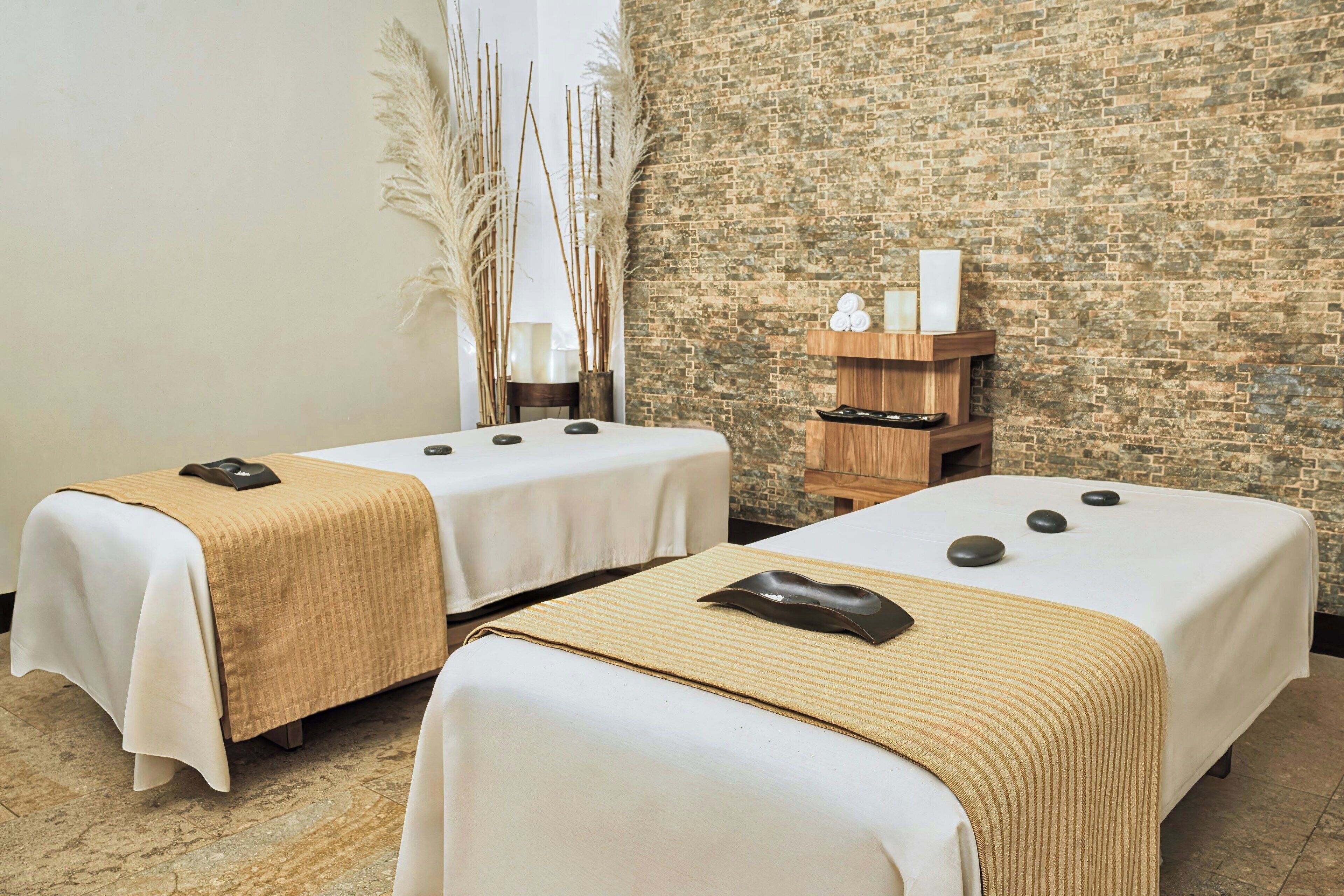 Deux tables de massage avec des pierres noires, une étagère en bois ornée d’objets décoratifs et de hautes herbes des pampas dans une salle de spa avec un mur en briques.