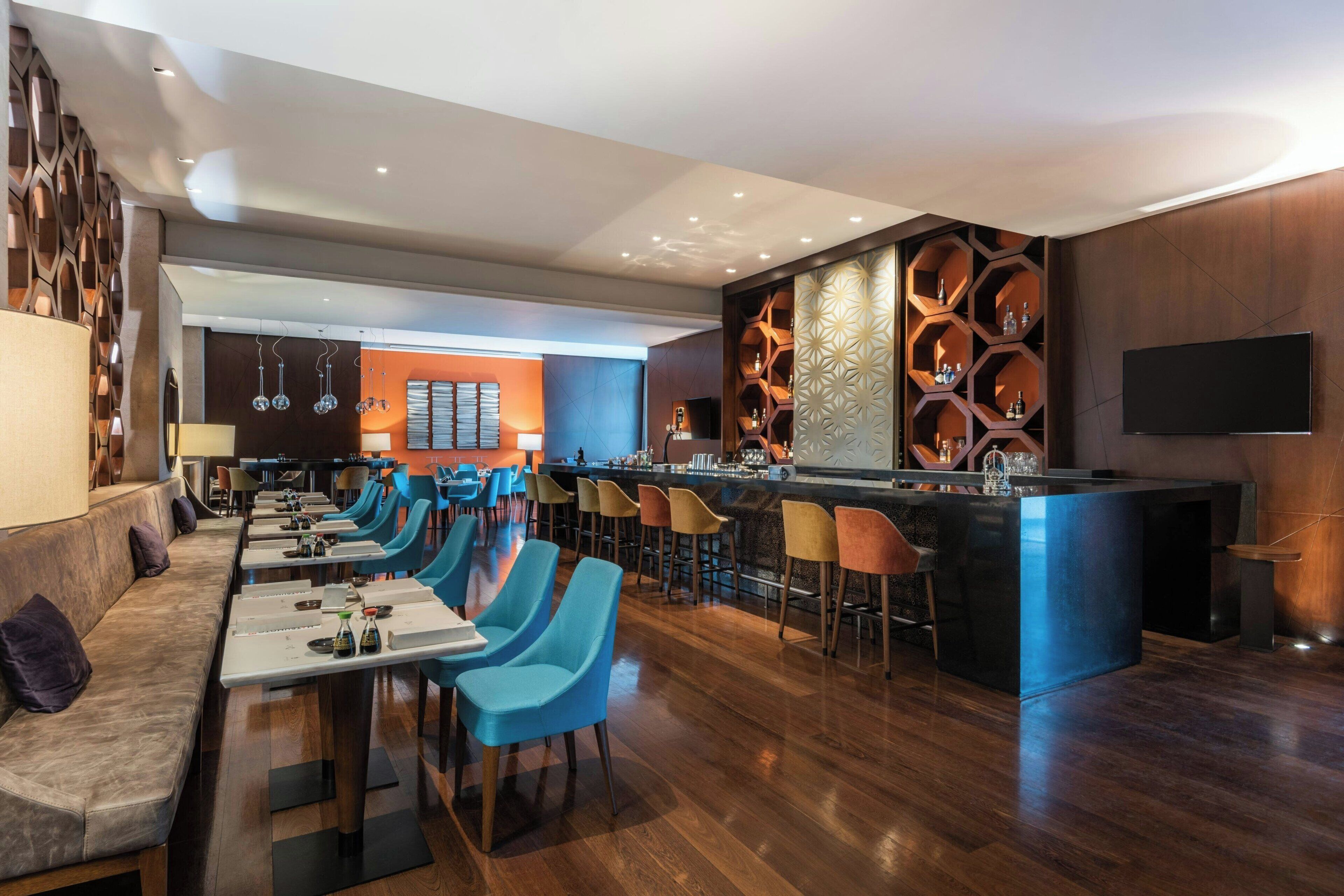 A modern bar area with a long counter, blue chairs, and a seating area with a sofa and tables.