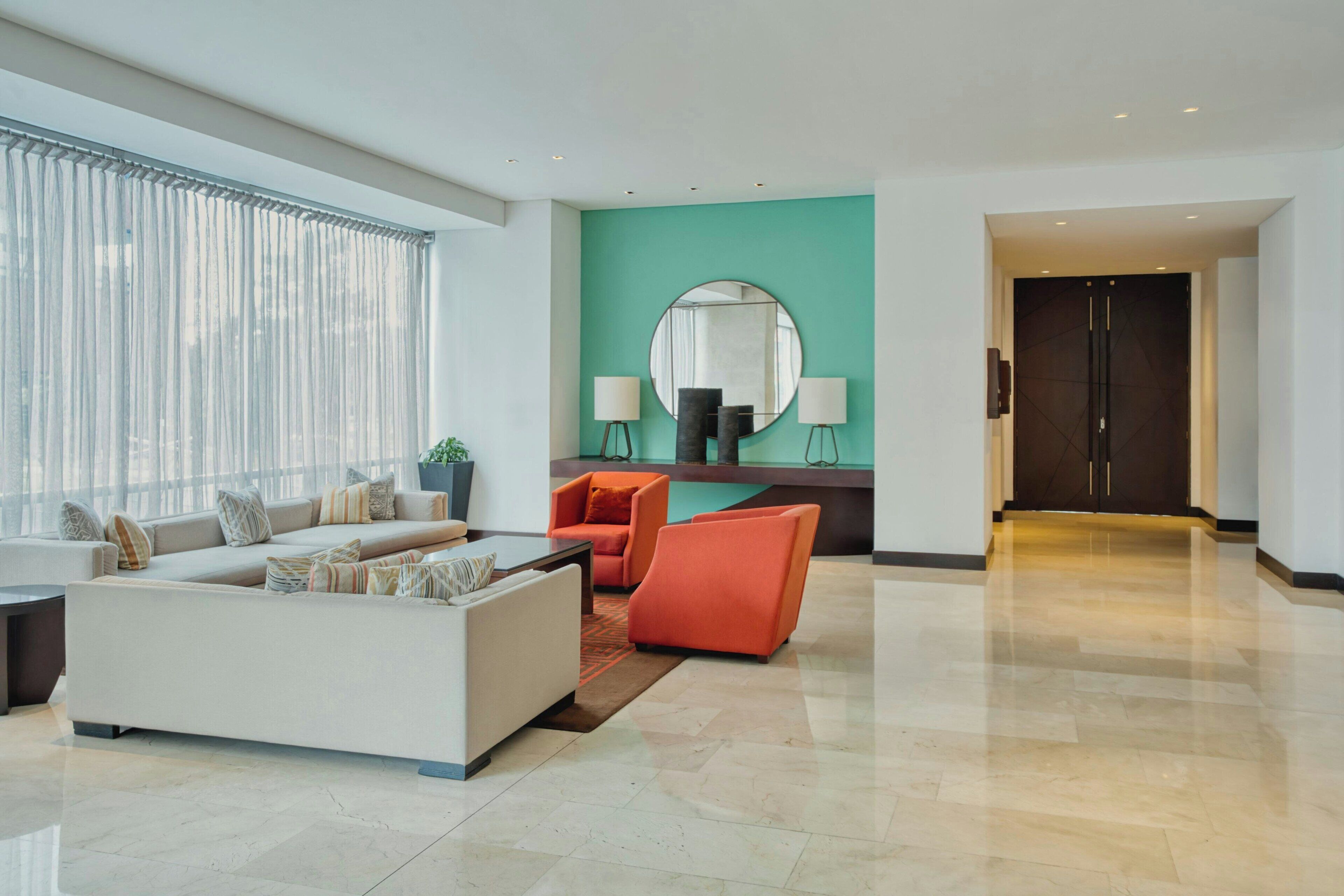 A modern living room with a sofa, armchairs, a coffee table, a large window with curtains, a round mirror, and a dark door.