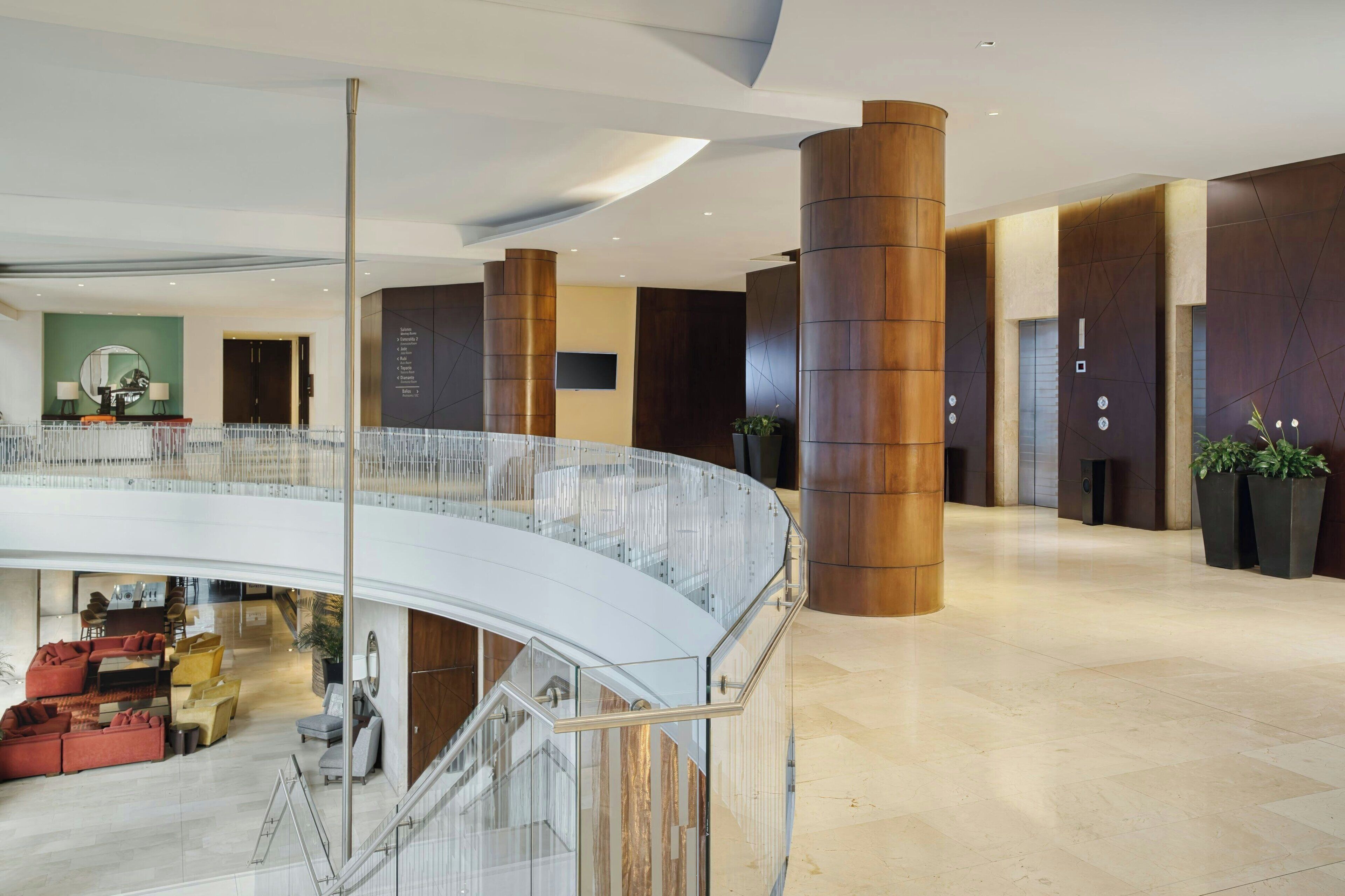 Eine moderne Hotelhalle mit gewölbtem Glasgeländer, einer zentralen Treppe und einer Sitzecke mit Sofas.