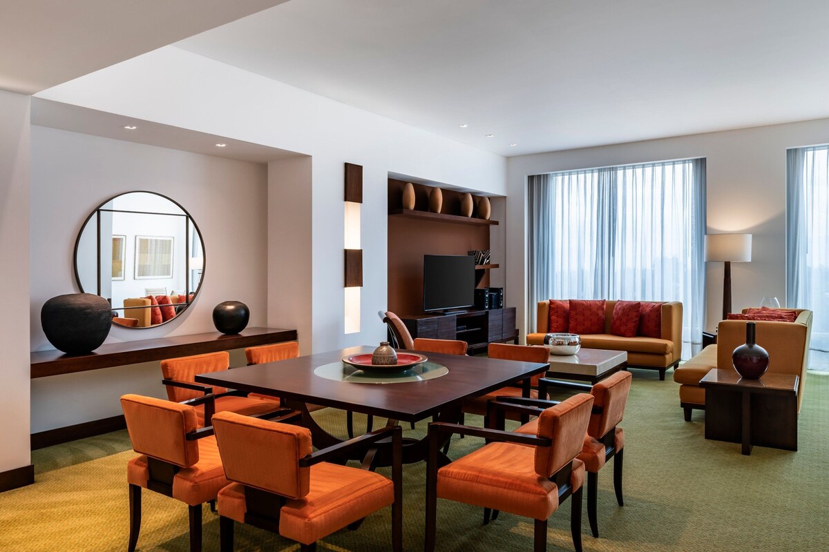 Una habitación de hotel moderna con comedor, zona de estar con sofá y sillones, televisor de pantalla plana y un amplio ventanal con cortinas.