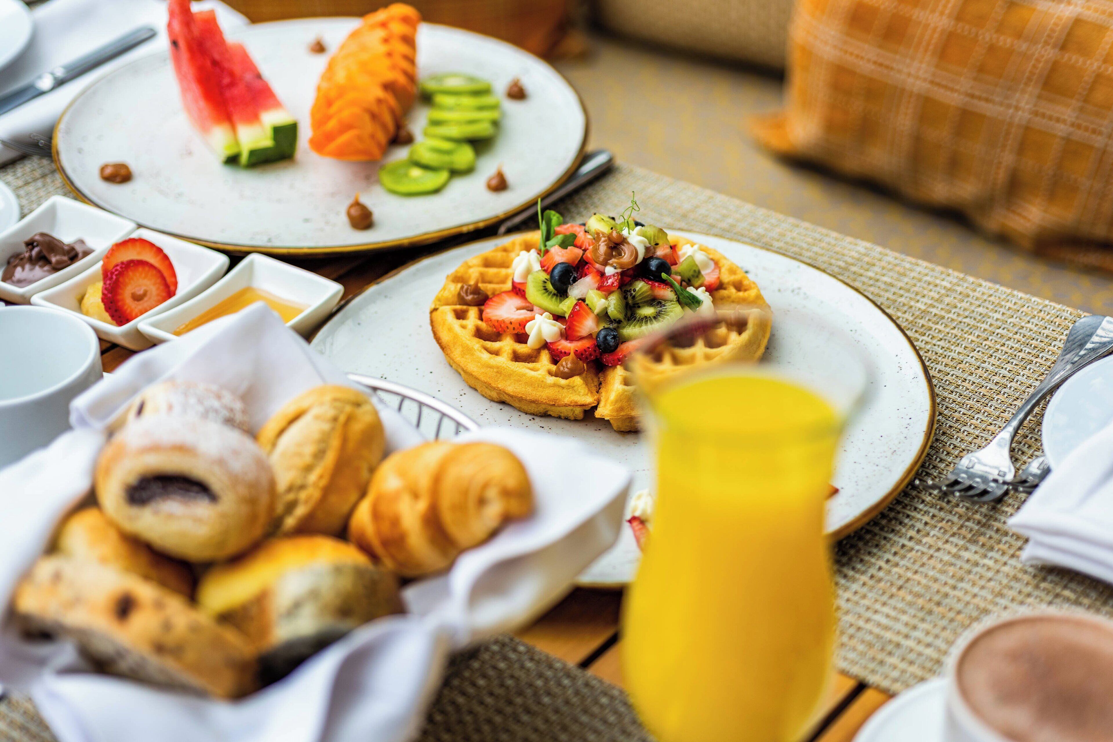 A breakfast setting with waffles topped with fruits and a basket of croissants.