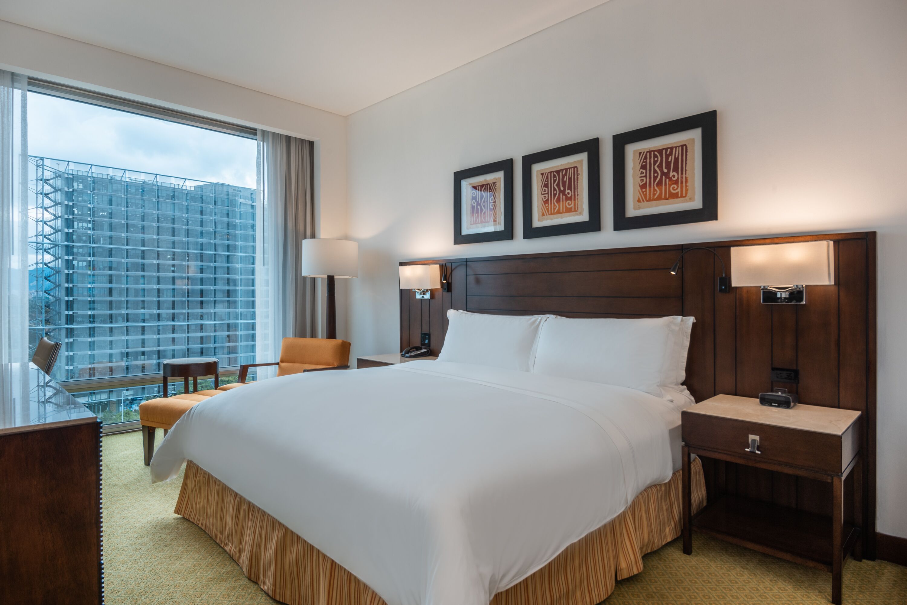 A hotel room with a large bed, two bedside tables, a desk, and a chair. There are three framed pictures on the wall and a view of a building outside the window.