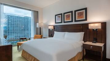 A hotel room with a large bed, two bedside tables, a desk, and a chair. There are three framed pictures on the wall and a view of a building outside the window.