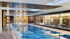 Una piscina interior moderna con paredes de vidrio transparente, techo de madera y un área de ejercicios visible.