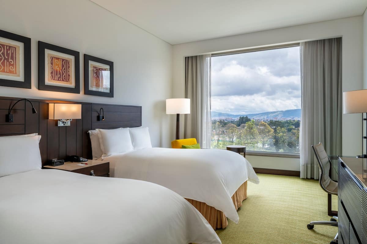 A hotel room with two beds, a desk, a chair, a TV, and a large window with a city view.