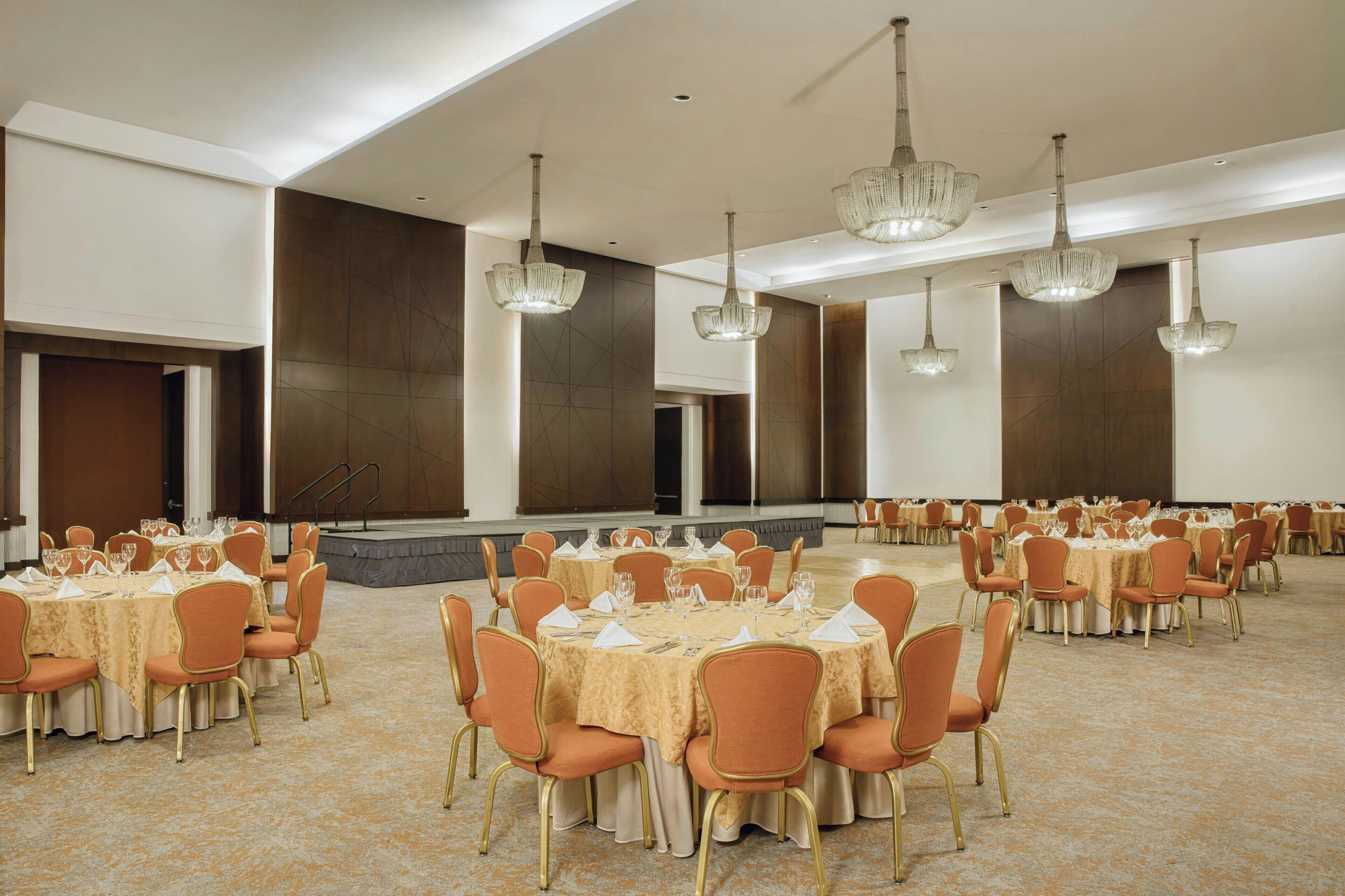A banquet hall with round tables, orange chairs, and chandeliers.