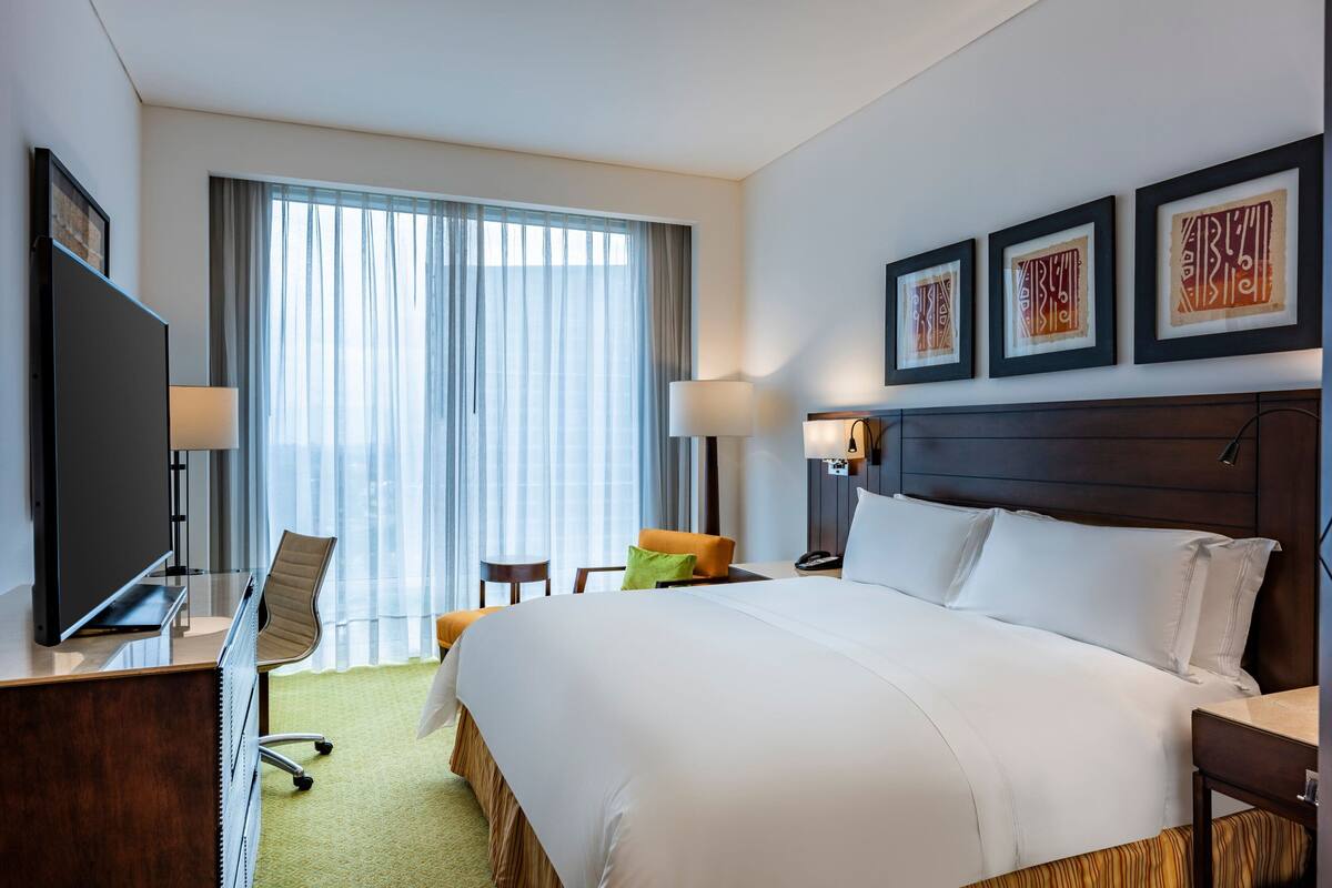 A hotel room with a large bed, a desk with a TV, a chair, a lamp, and a window with curtains.