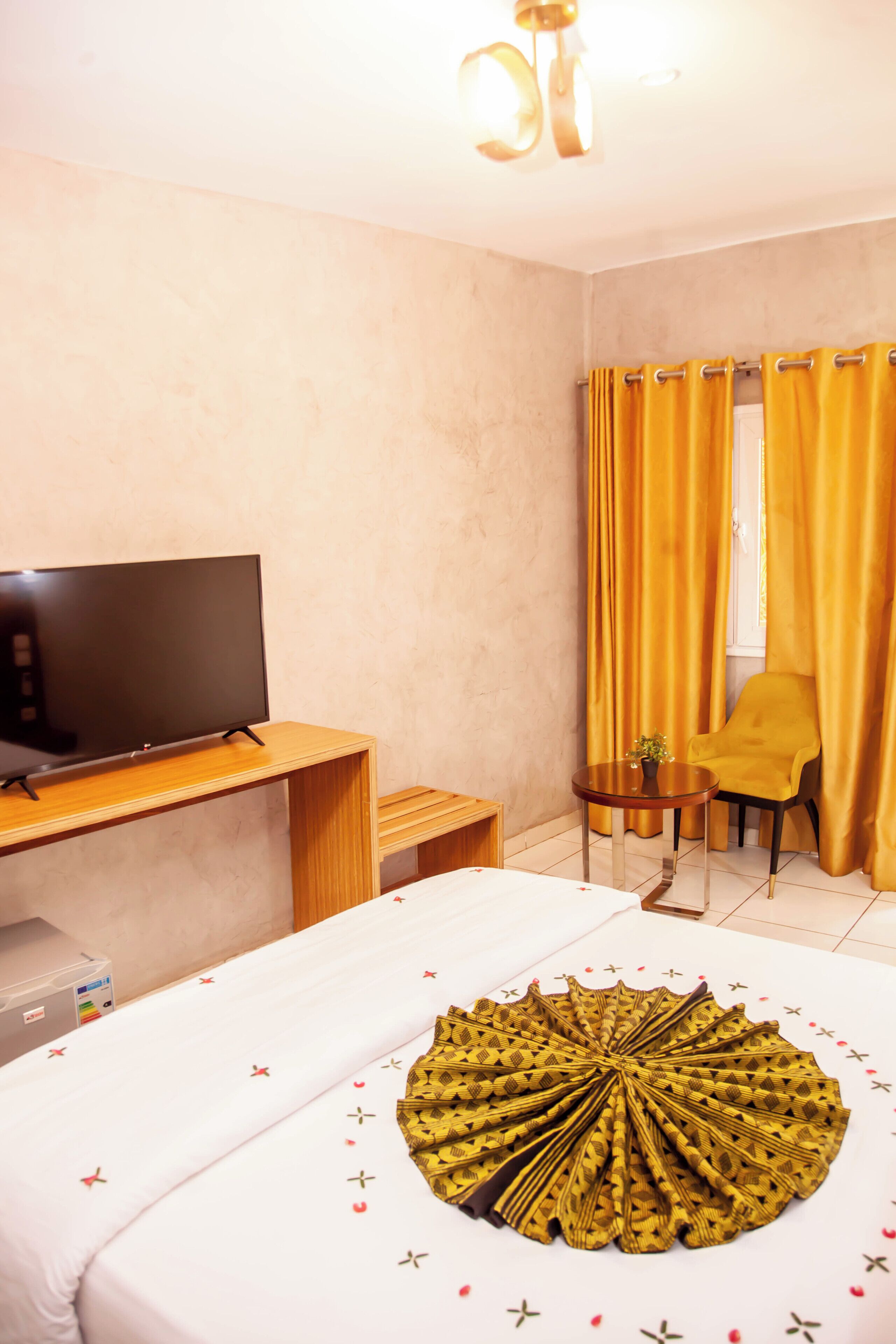 A hotel room with a bed, a TV on a wooden stand, a small round table with a chair, and yellow curtains.