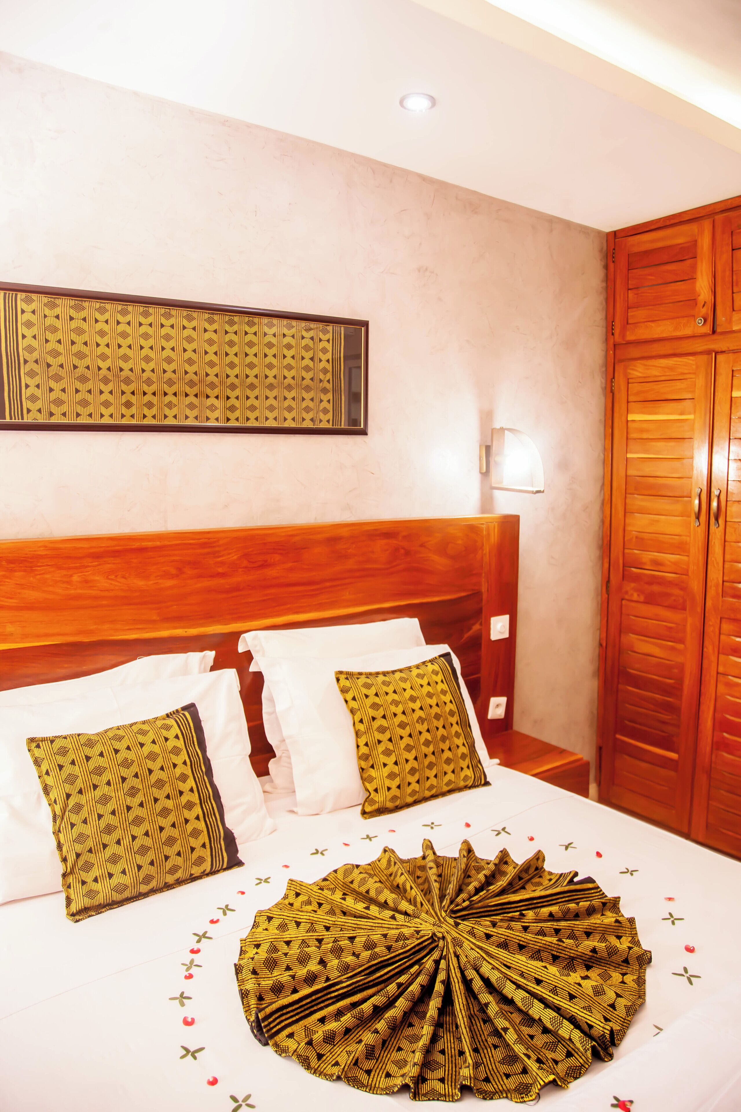 A hotel room with a wooden headboard, a large bed with patterned pillows, a decorative fan, and a wardrobe.