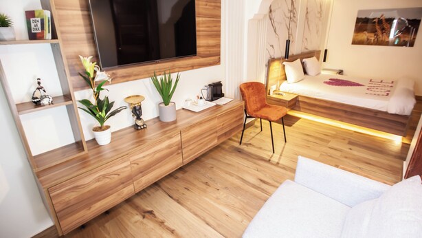 A modern bedroom with a large TV mounted on a wooden wall, a bed with a headboard, and a seating area with a chair and a sofa. - Hotel Flamboyants (Mbour)