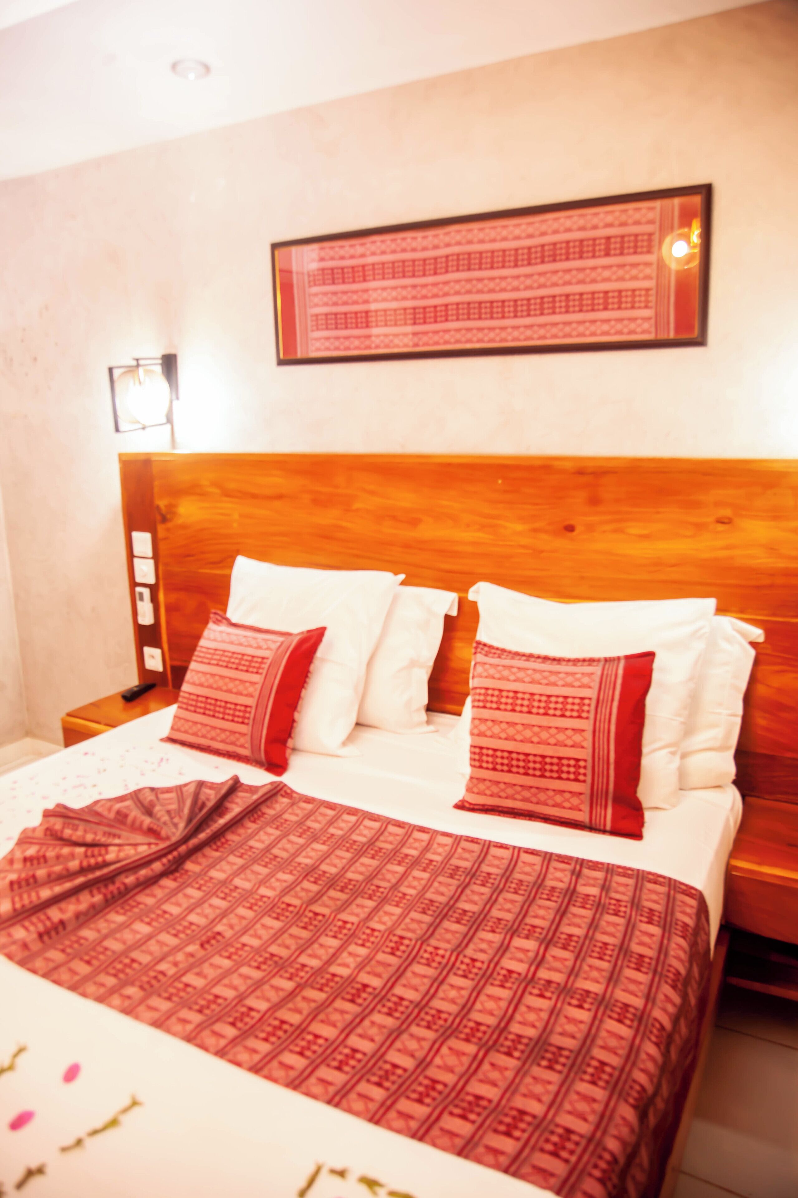 A hotel room with a wooden headboard, a bed with white and red bedding, a framed picture above the bed, and a nightstand with a lamp and a remote control.