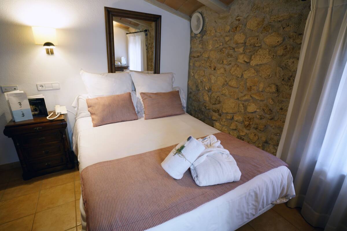 A hotel room with a stone wall, a bed with white and pink bedding, a nightstand with a phone and a brochure, and a mirror above the bed.