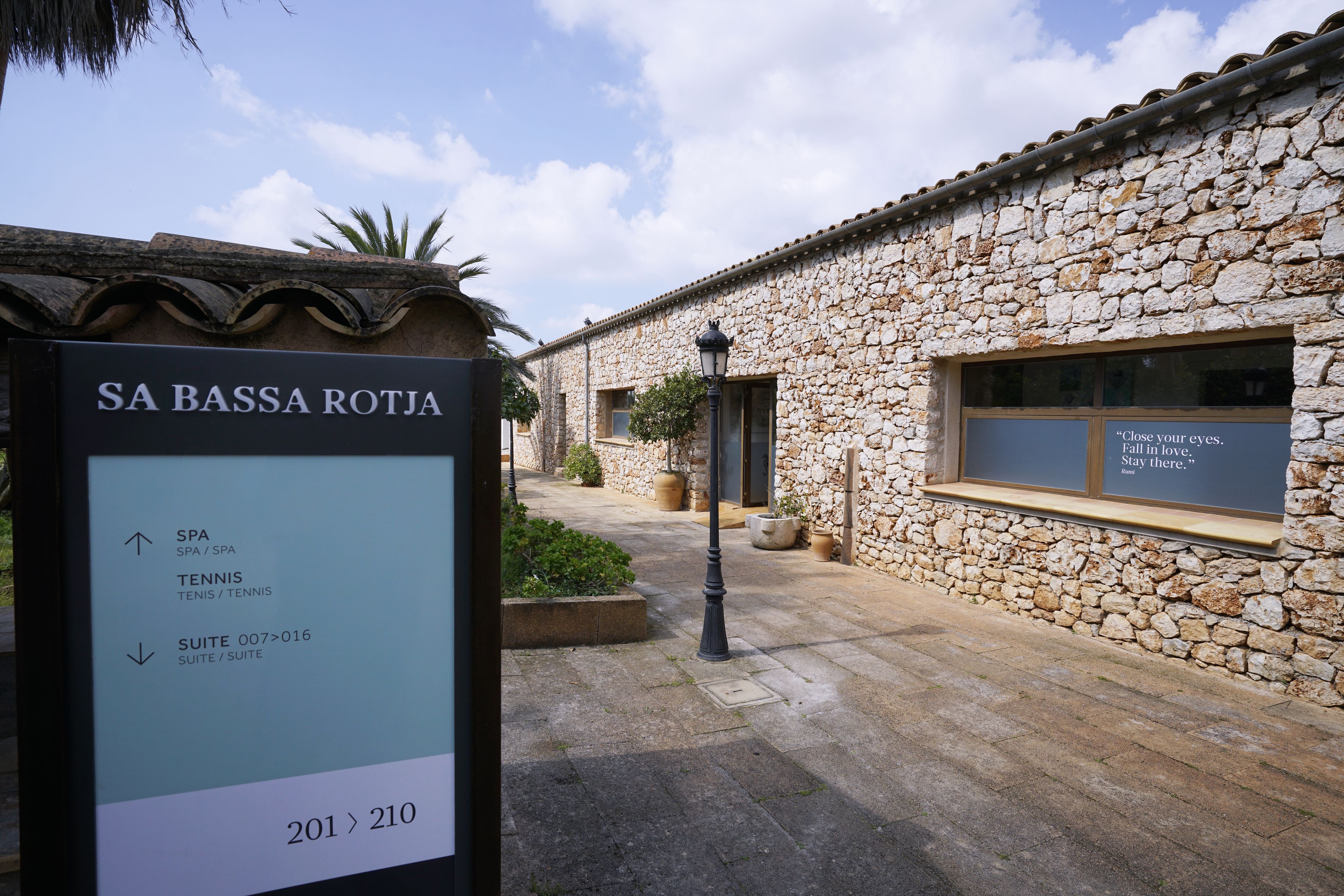 Een stenen gebouw met een bord waarop 'SA BASSA ROTJA' staat, en voorzieningen zoals een SPA, TENNIS en SUITE 007>016.