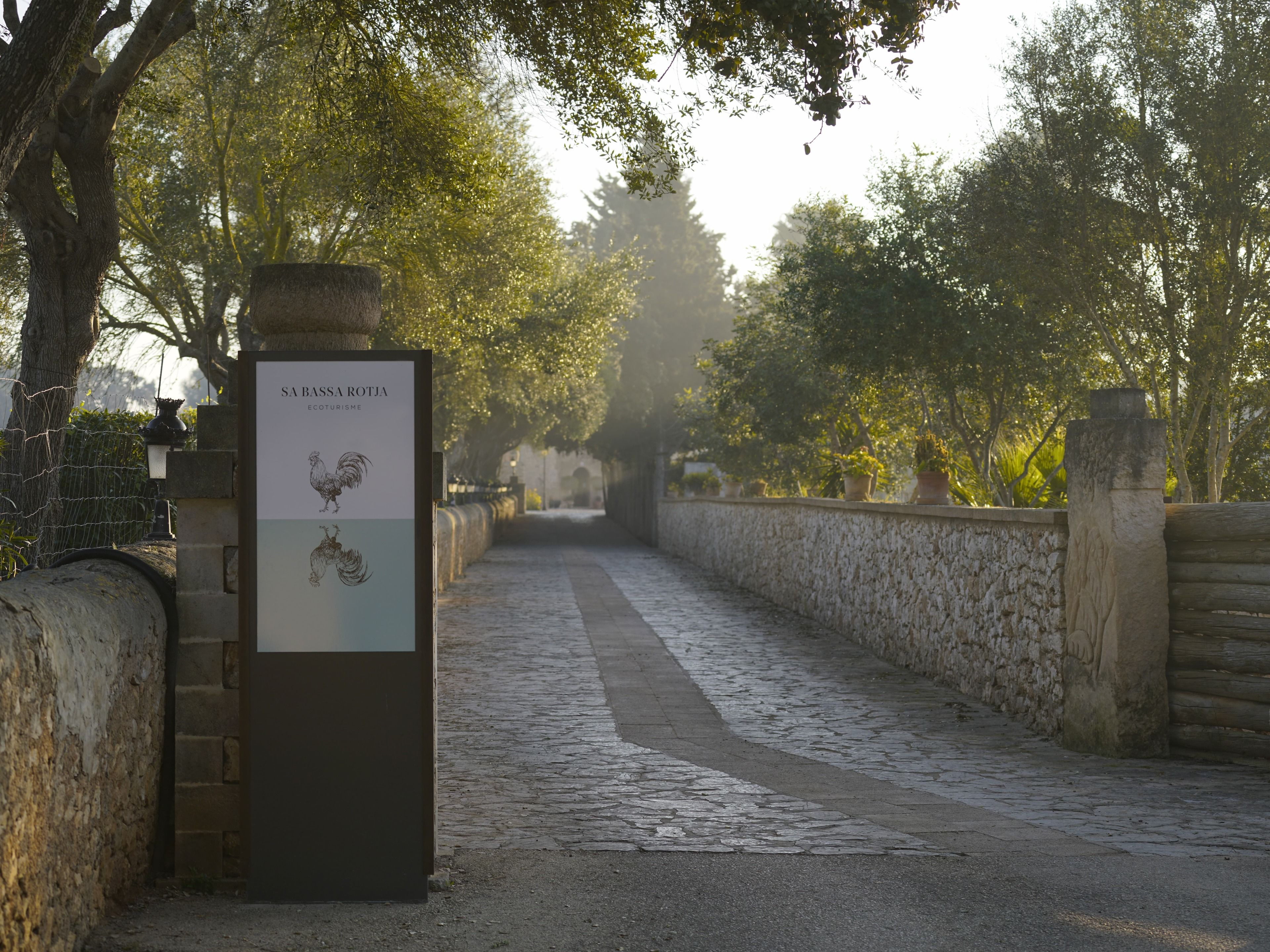 A cobblestone pathway flanked by stone walls and trees, leading to a distant structure. A signboard with text and illustrations is visible on the left.