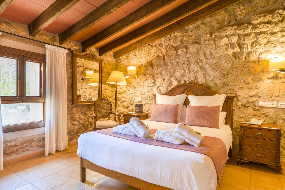 A bedroom with a stone wall, wooden bed frame, two bedside lamps, a chair, a nightstand, and a window with curtains.