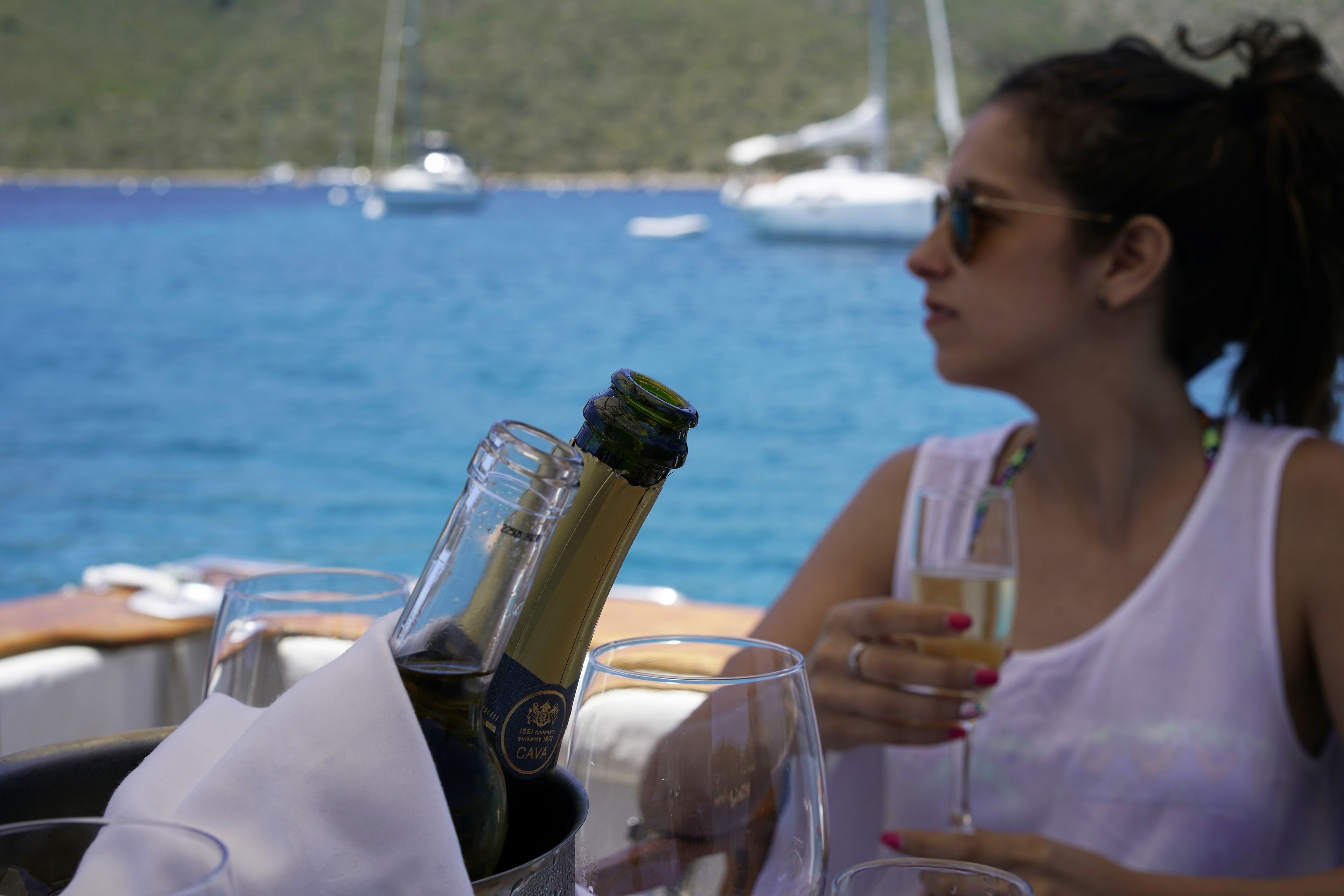 Una mujer sentada a una mesa con una botella de champán y copas, con vistas a una masa de agua donde se ven barcos al fondo.