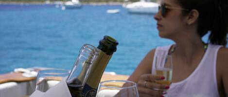 A woman sitting at a table with a bottle of champagne and glasses, overlooking a body of water with boats in the background.