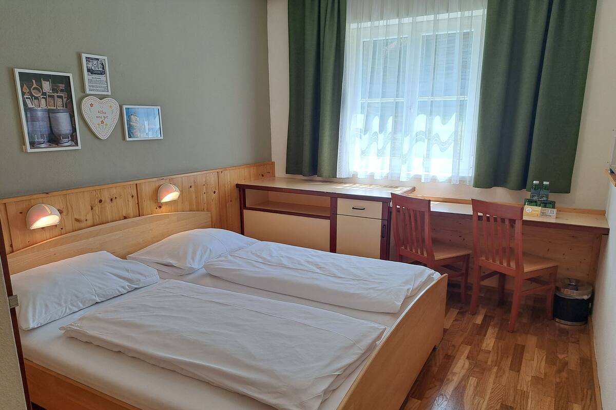 Een kamer met een houten bed, een bureau en een stoel. Er hangen foto's aan de muur en er is een raam met gordijnen.