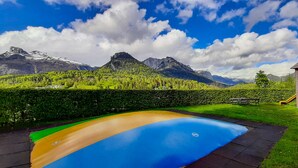 Una piscina con un tobogán de colores, una estructura de juego de madera y vistas a montañas bajo un cielo azul.