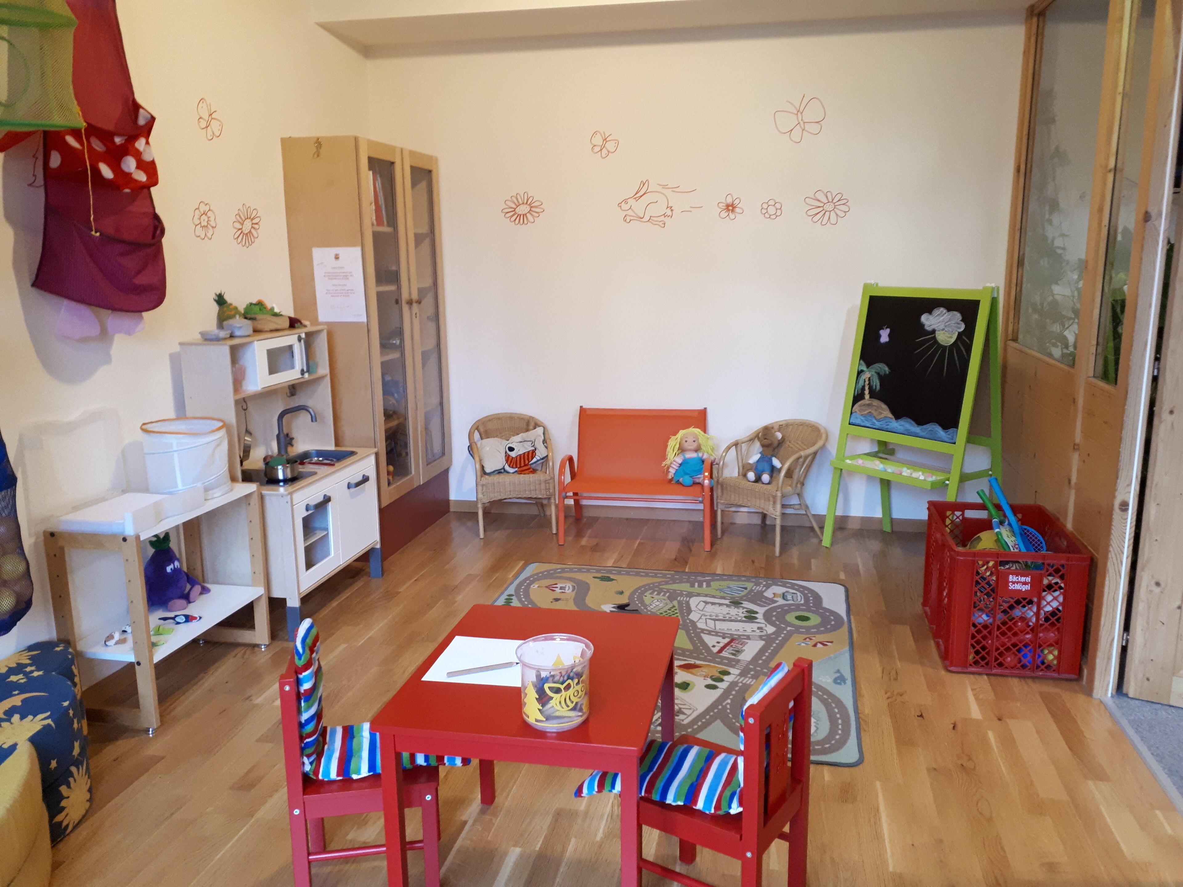 A children's playroom with a red table, two red chairs, a play mat, a bookshelf, a small cabinet, and a toy storage bin.
