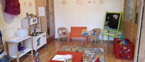A children's playroom with a red table, two red chairs, a play mat, a bookshelf, a small cabinet, and a toy storage bin.