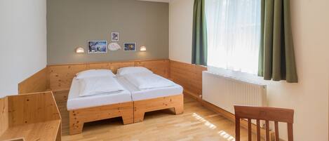 Una habitación con una cama de madera, un escritorio y una silla. Se observa una ventana con cortinas, un radiador y luces fijadas a la pared.