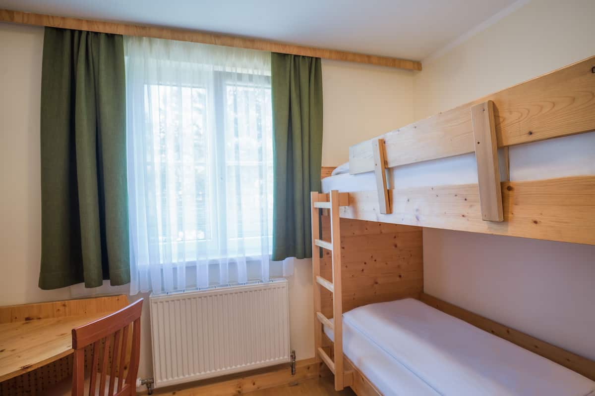 Een slaapkamer met een stapelbed, een houten bureau en stoel, een raam met gordijnen en een radiator.