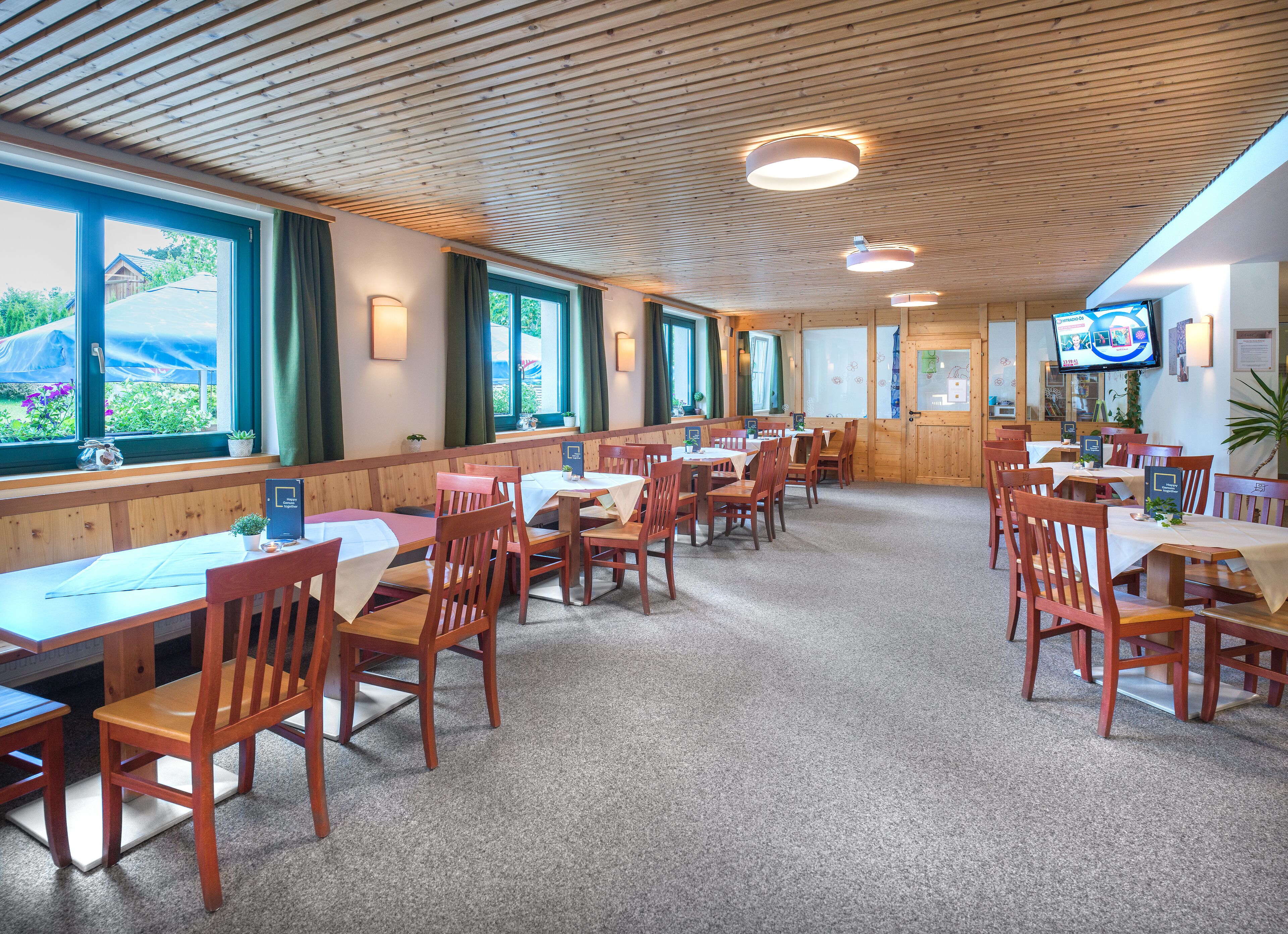 A restaurant with wooden tables and chairs, large windows, and a television on the wall.