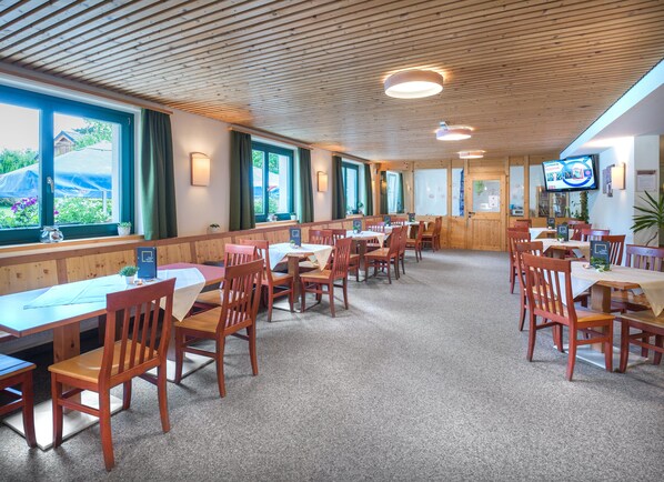 A restaurant with wooden tables and chairs, large windows, and a television on the wall. - JUFA Hotel Bad Aussee (Bad Aussee)
