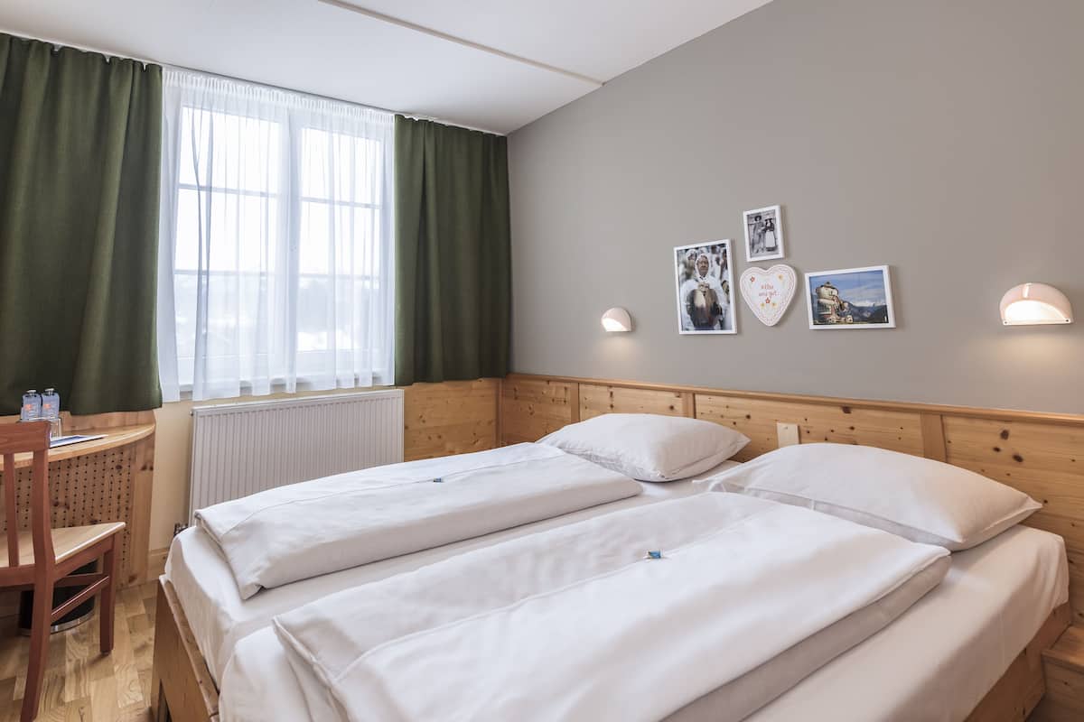 Een kamer met twee bedden, een houten hoofdbord, een raam met groene gordijnen, een radiator en aan de muur hangende foto's.