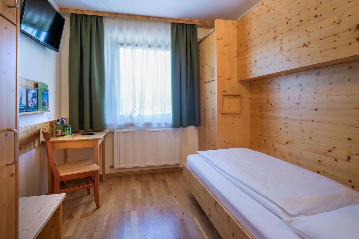 Een kleine kamer met een bed, bureau, stoel, een aan de muur bevestigde televisie en een raam met gordijnen.