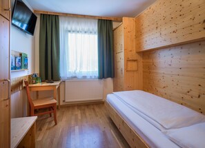 A compact room with a bed, a desk, a chair, a TV mounted on the wall, and a window with curtains.