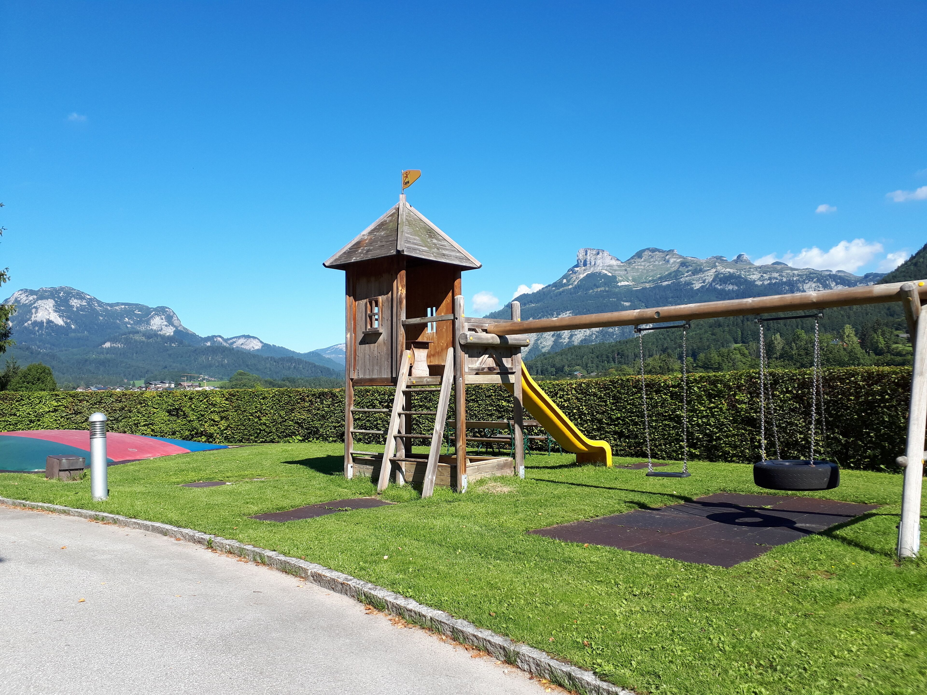 A playground with a wooden playhouse, a yellow slide, and a swing set, set against a backdrop of mountains and clear blue sky.