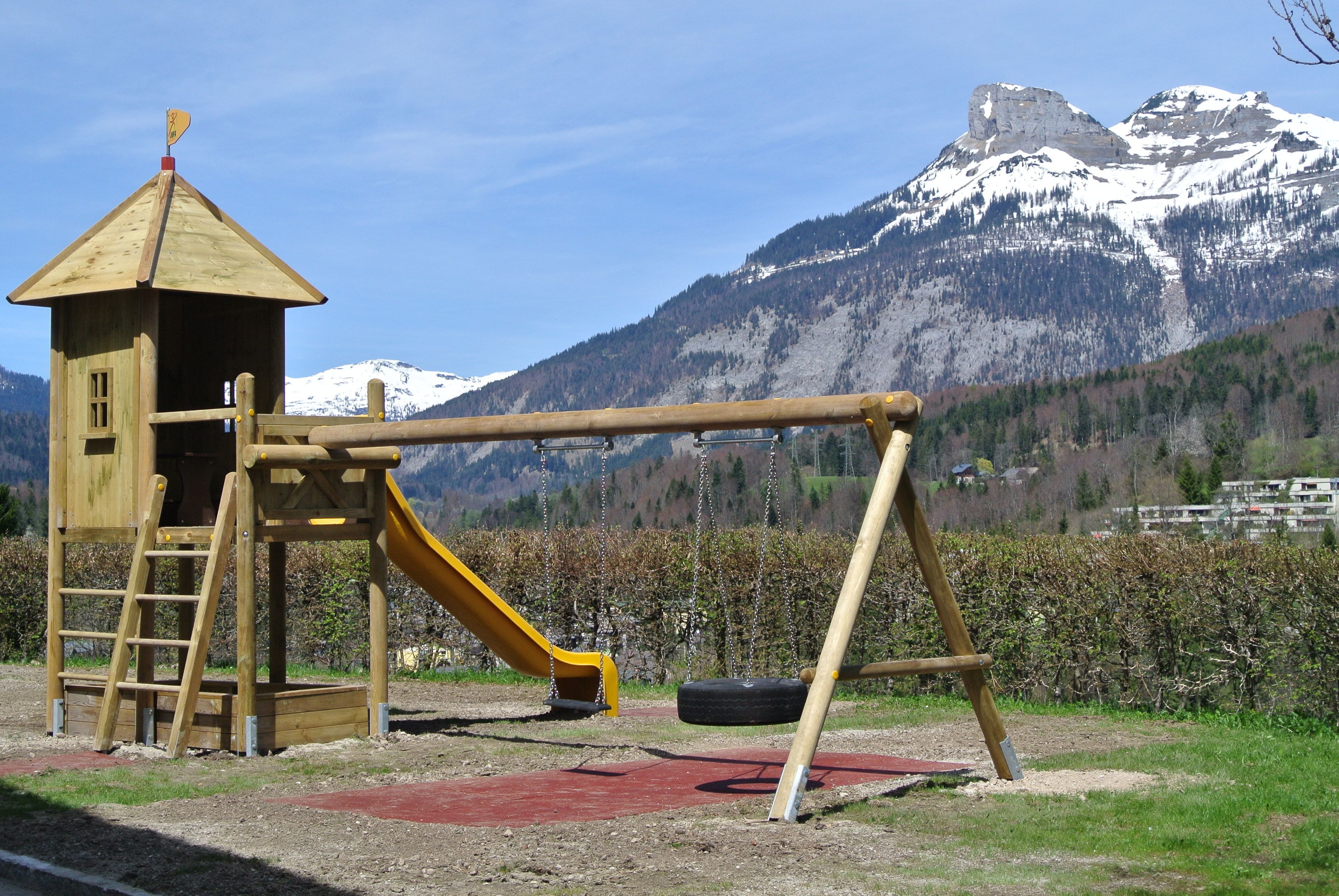 A playground with a wooden slide and swing set, set against a backdrop of snow-capped mountains.