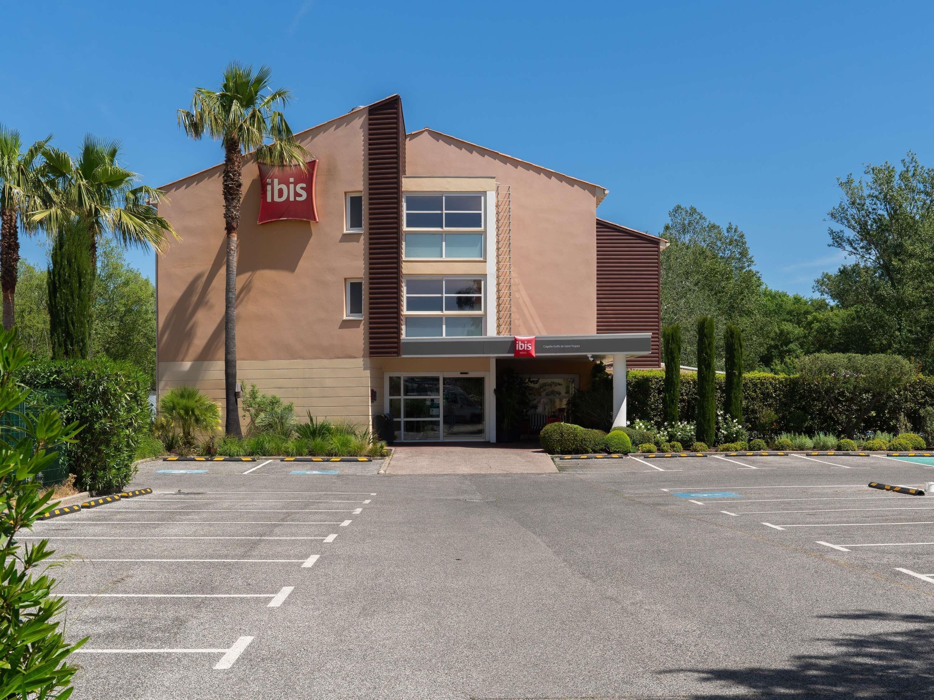A modern building with a 'ibis' sign, a parking lot, and surrounding greenery.