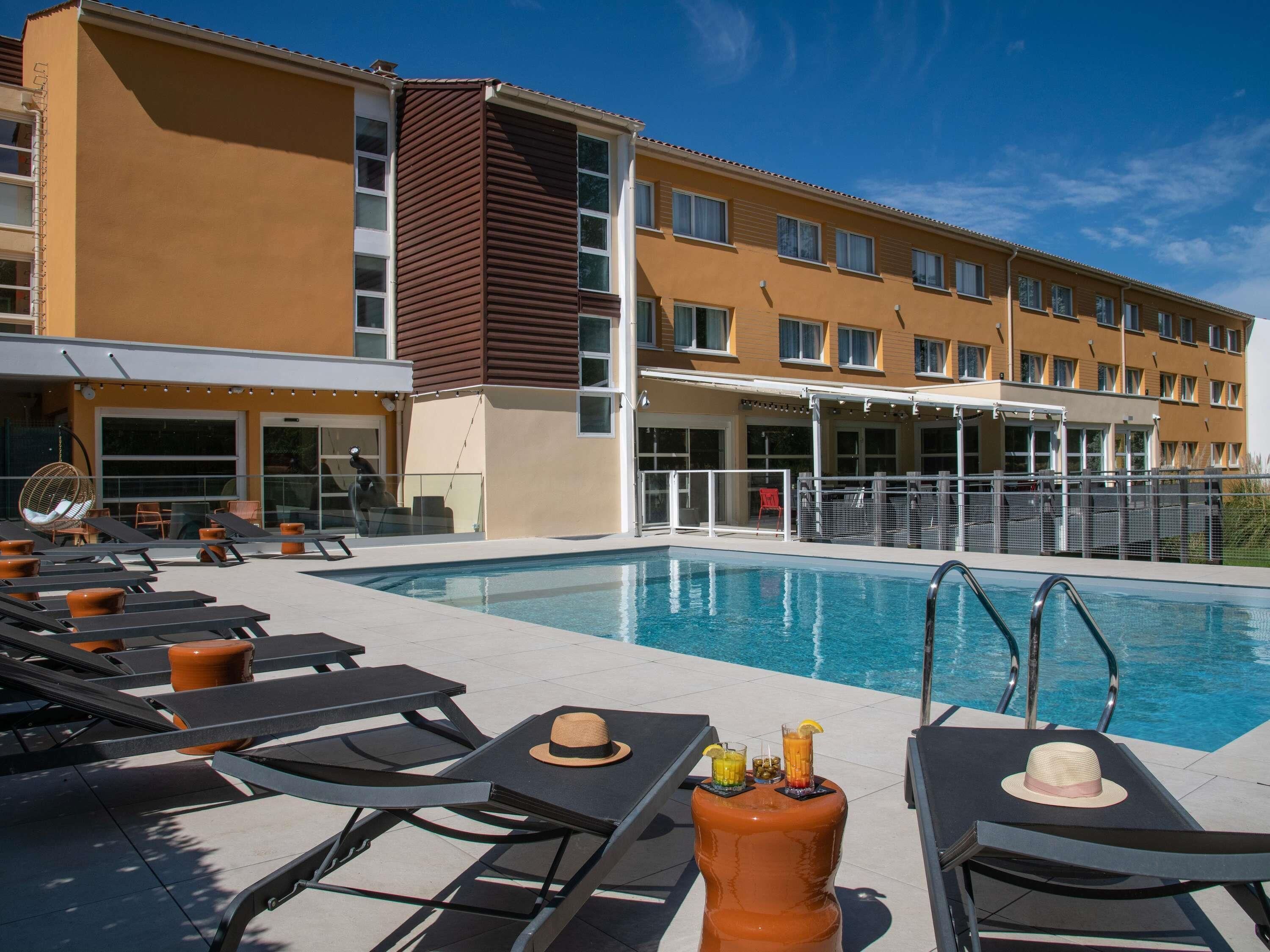 A hotel pool area with sun loungers, a large pool, and a building with multiple windows.