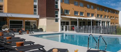 A hotel pool area with sun loungers, a large pool, and a building with multiple windows.