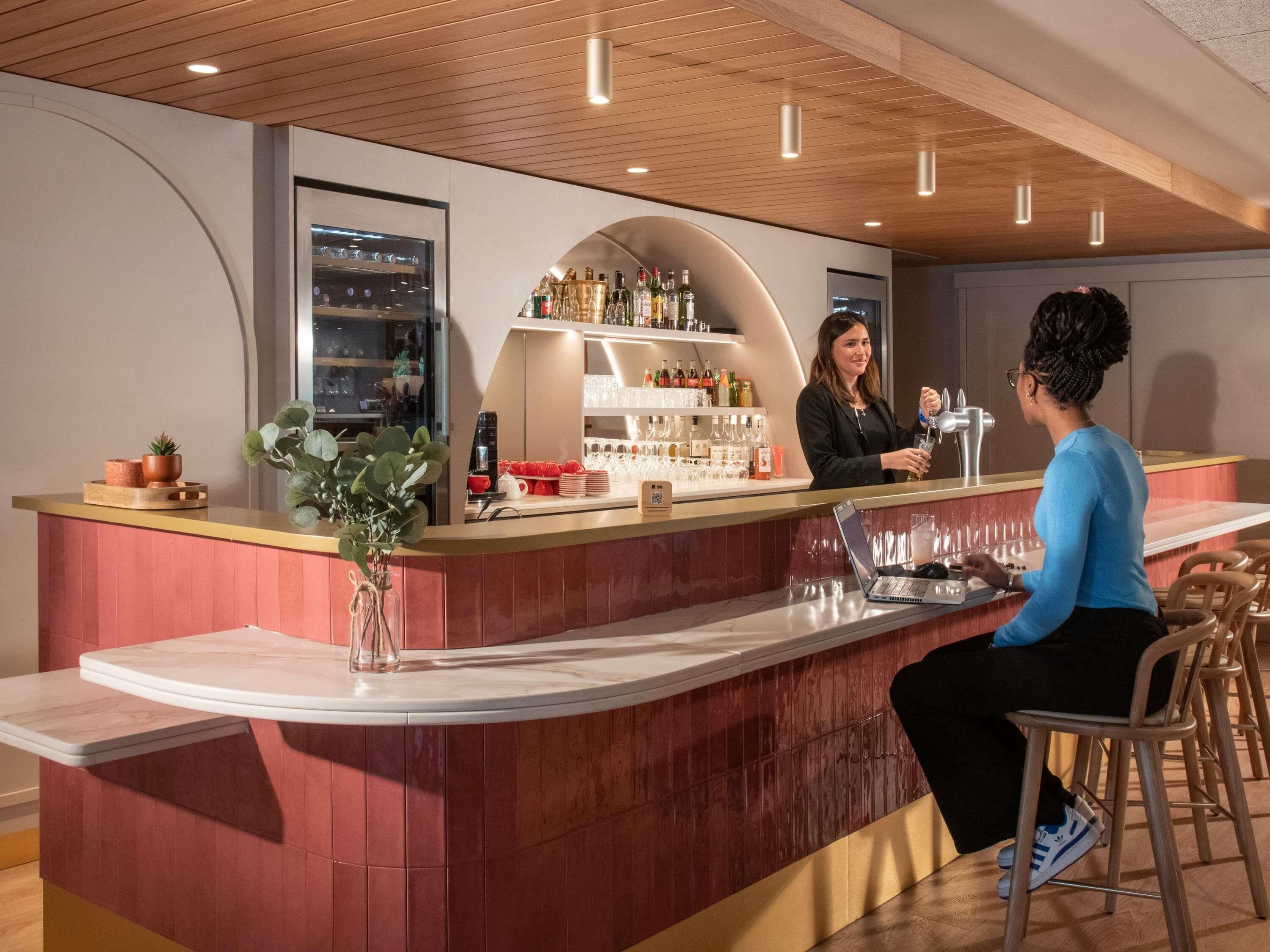 Un área de bar moderna con una barra curva, una computadora portátil y un barista preparando una bebida.
